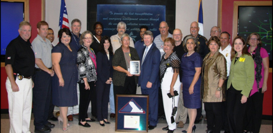 Group of officials and community members standing together indoors, posing for a photo during an award presentation, with flags and a screen in the background.